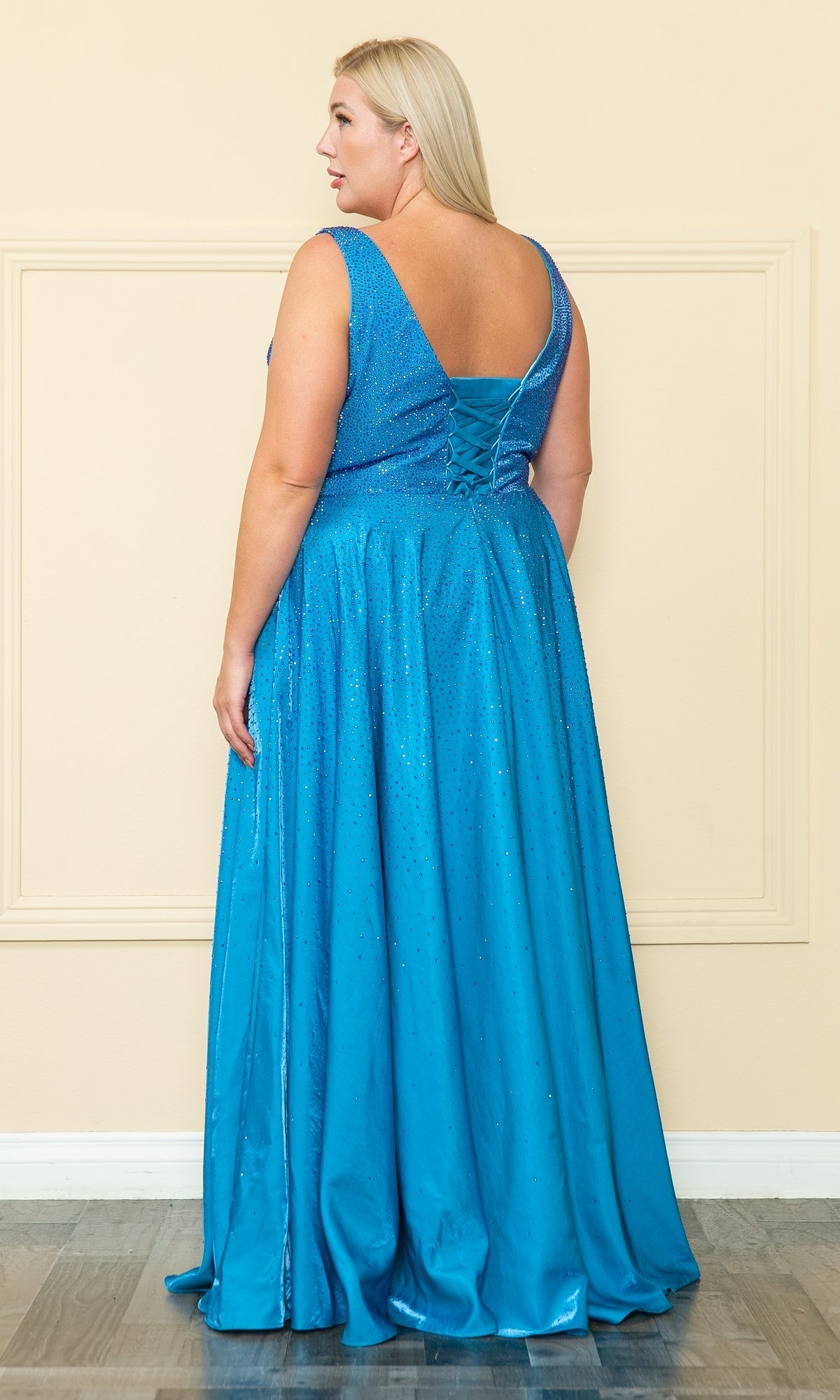 A woman with long blonde hair wears the Embellished Plus-Size Long V-Neck Prom Dress W1110—a sleeveless, floor-length blue gown with a low back and lace-up detail—standing on a wooden floor against a cream-colored wall.